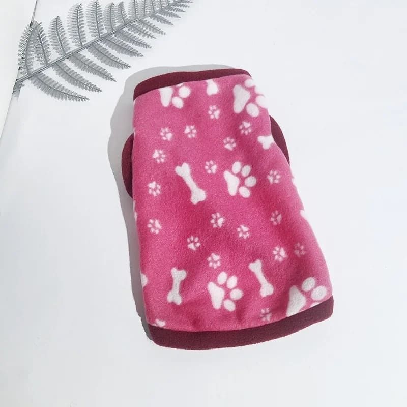 A pink fleece jacket for pooches featuring a pattern of white paw prints and bones, finished with a dark trim, presented on a bright background with a delicate fern leaf to the side.