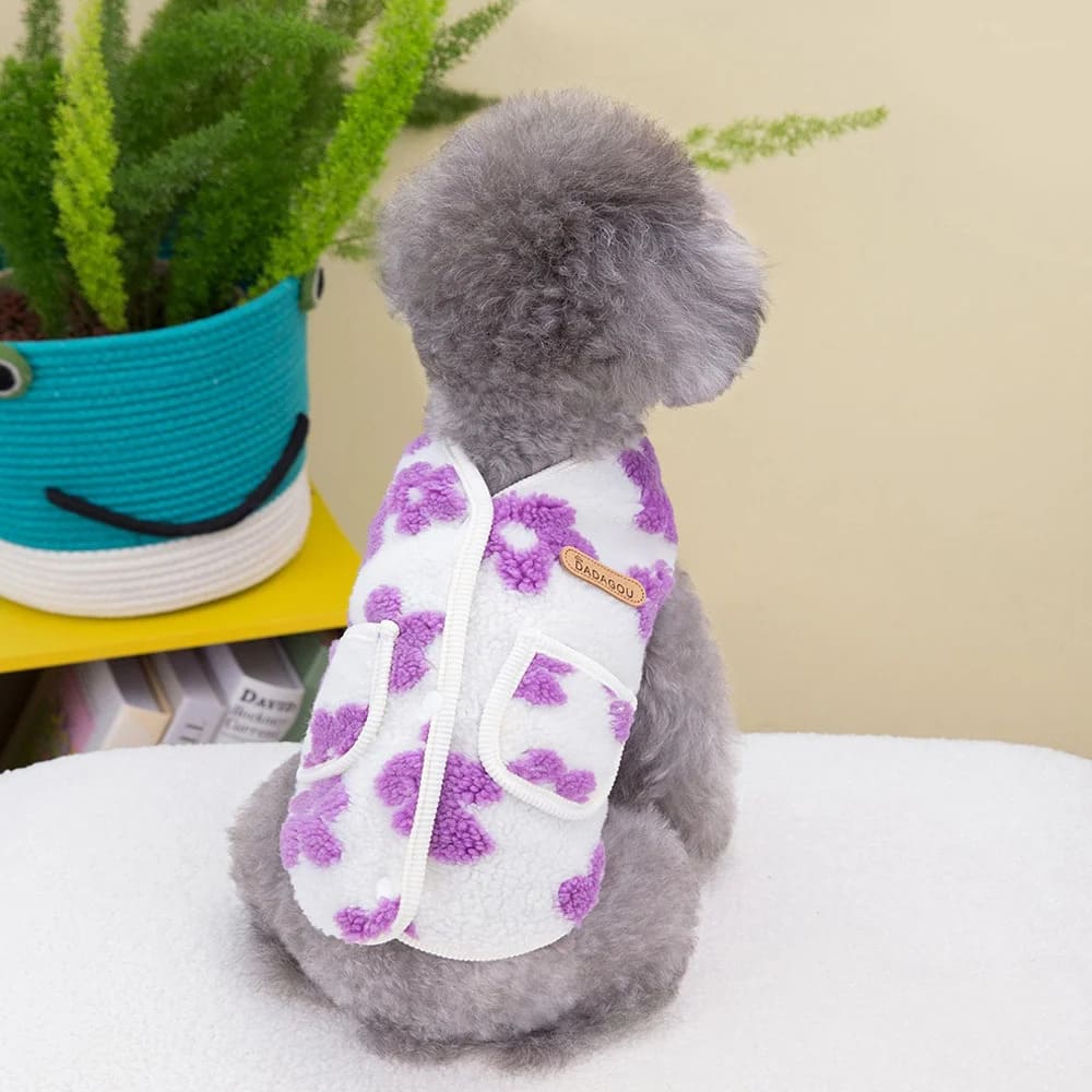 The image shows a charming little dog wearing a purple floral fleece vest. The vest features a white base with lovely purple flowers scattered across it, and it secures with what appears to be a Velcro fastening system along the dog's back.